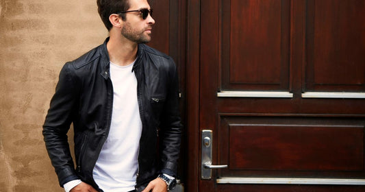 A man standing next to a door wearing a leather jacket, white tshirt, sunglasses, and watch.