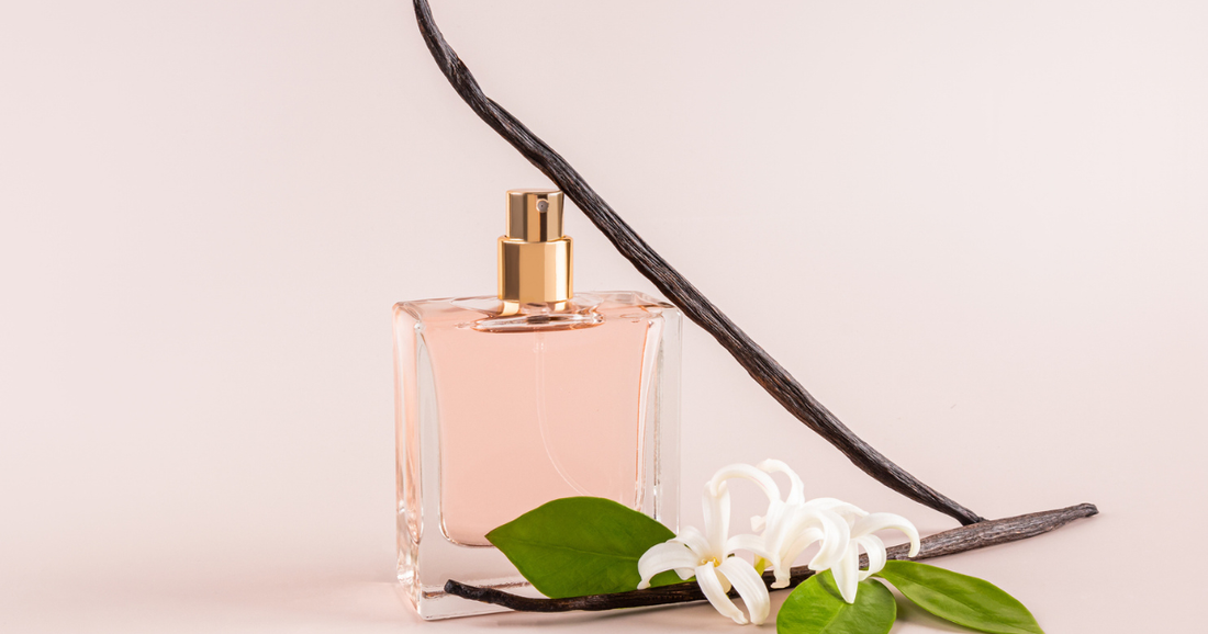 A pink perfume bottle with two vanilla beans and a vanilla bean flower against a light pink background.