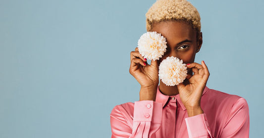 A black woman with short hair in a pink shirt holding two flowers in front of her face.